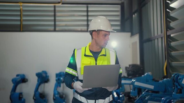 Worker reviews data on laptop near industrial robots. He wears safety gear and helmet with focused attention. Blue robots stand idle in background