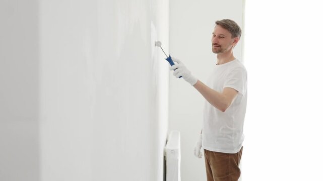Middle aged man wearing white gloves and t-shirt, is painting white wall with paint roller, experiencing home renovation and transforming living space