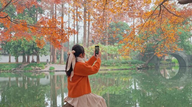Female traveler holding smartphone taking pictures of a classic wooden boat on calm lake surrounded by autumn foliage in Hangzhou, China