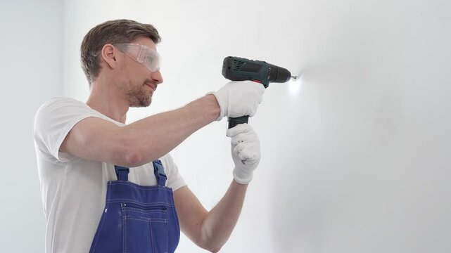 Professional handyman wearing blue overall, safety glasses and gloves, is using cordless drill to make hole in white wall during renovation or installation work