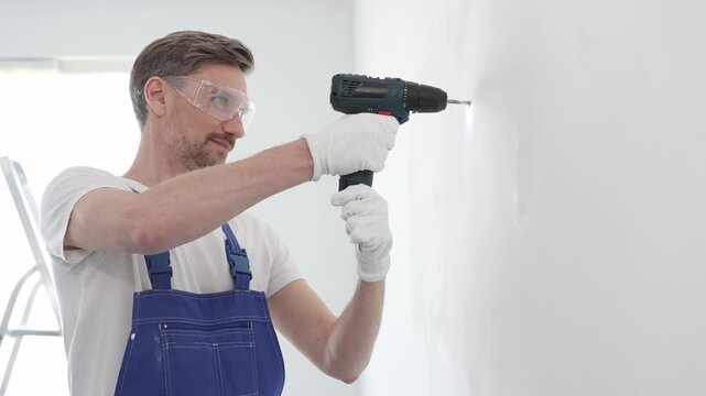 Professional handyman wearing blue overall, safety glasses and gloves, is using cordless drill to make hole in white wall during renovation or installation work