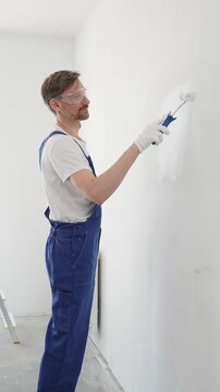 Professional painter wearing protective white gloves and blue work overalls, applying paint with roller on pristine interior wall during home renovation project