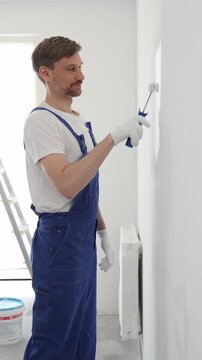 Professional painter wearing protective white gloves and blue work overalls, applying paint with roller on pristine interior wall during home renovation project