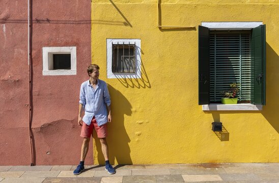 Young man leaning against a house wall, red and yellow house facade with windows, colourful houses on the island of Burano, Venice, Veneto, Italy