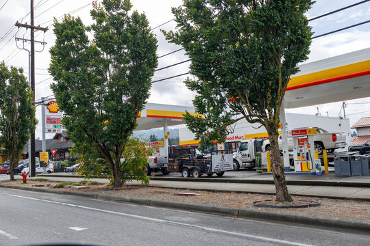 A Shell fuel station and food mart seen across a tree lined street, with work trucks and a utility trailer parked beneath the canopy on an overcast late morning.