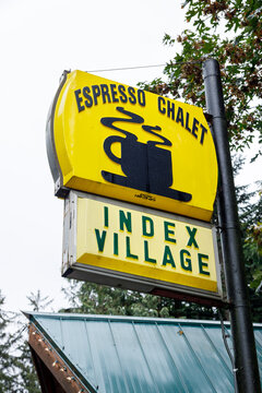 Looking up at the yellow Espresso Chalet sign and its steaming coffee cup logo above green Index Village lettering, the cafe green roof and evergreen branches below a pale sky.