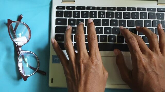 Top View of Hands Typing on a Laptop Keyboard with Eyeglasses on a Blue Desk