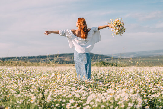 Joyful woman standing with arms outstretched holding a bouquet of Matricaria chamomilla in a sunlit meadow