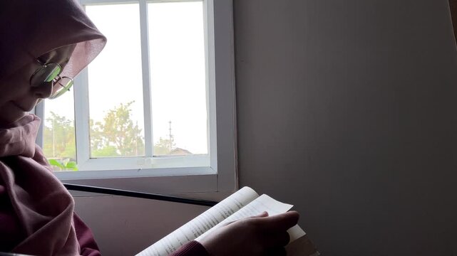 Slow motion low angle side view of an Asian Muslim woman in a hijab and glasses. She is sitting indoors reading a book by a bright window, enjoying a quiet and peaceful study time at home.