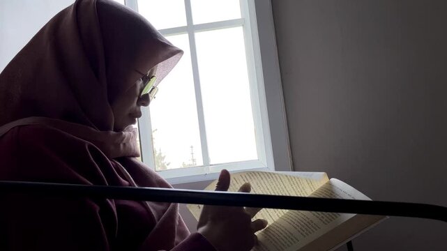 Slow motion worm's eye view of an Asian Muslim woman wearing a hijab and glasses. She is sitting indoors reading a book by a bright window, enjoying a quiet and peaceful leisure time at home.
