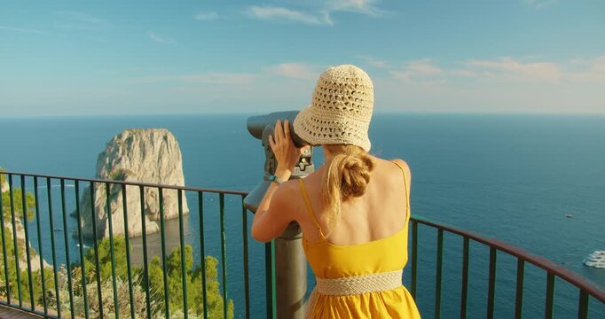 Woman sightseeing with binoculars on a Capri terrace above Faraglioni sea cliffs on a summer day