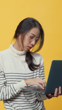 Vertical Screen : Unhappy Asia lady worker with casual cloth holding and typing laptop computer scratch her hair displeased upset angry headache with overwork isolated on the yellow background.