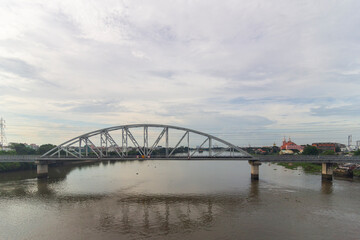 Obraz premium A steel arch bridge and elevated roadway cross an urban river beneath a broad cloudy sky with power lines and city buildings in the background.