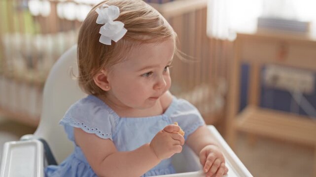 Toddler girl holding a small cracker snack with hand to mouth, wearing white bow, seated in highchair inside building; gentle curiosity.