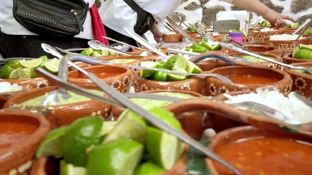 Mexican food buffet featuring decorated clay bowls filled with sauces, fresh lime wedges, serving utensils, and people preparing dishes at a food service in Mexico.