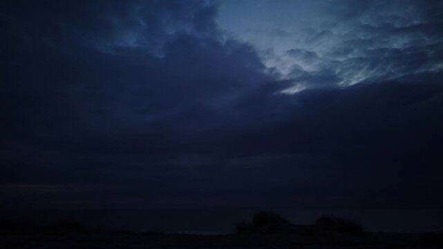 Wide landscape time-lapse from pitch darkness to dawn over sea with fast-moving dark overcast clouds revealing soft pink sunrise light on horizon.