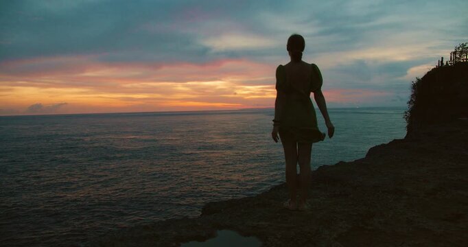 Woman silhouette standing on cliff edge watching warm colors of sunset reflecting on ocean surface