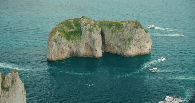 Rocky islet of Faraglioni sea stacks with boats circling in deep blue waters on sunny holiday day