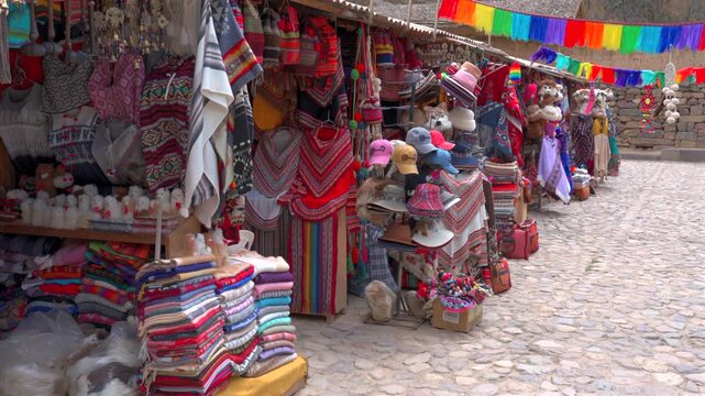 Wide shot of traditional artisan market stall displaying colorful alpaca textiles, ponchos, and souvenirs along a cobblestone street in Ollantaytambo, Sacred Valley, Cusco, Peru.