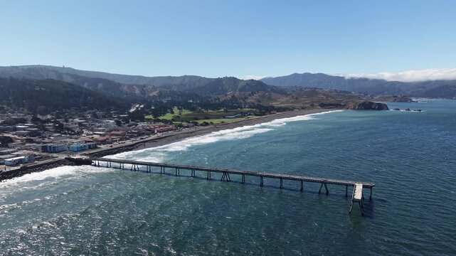 Aerial drone footage flying over the Pacifica Municipal Pier and the scenic California coastline, showcasing the town, sandy beach, and rolling hills under a clear blue sky on a sunny day