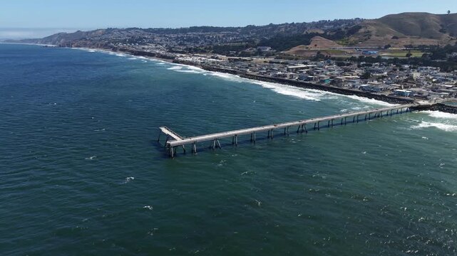 Aerial view of the Pacifica Municipal Pier and the coastline of Pacifica, California, showing the beautiful blue ocean water and gentle waves crashing on the shore on a sunny day