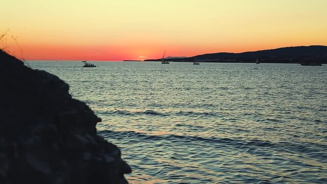 Vibrant sunset casts orange and purple hues over calm sea, with silhouetted boats and distant hills creating a tranquil coastal scene
