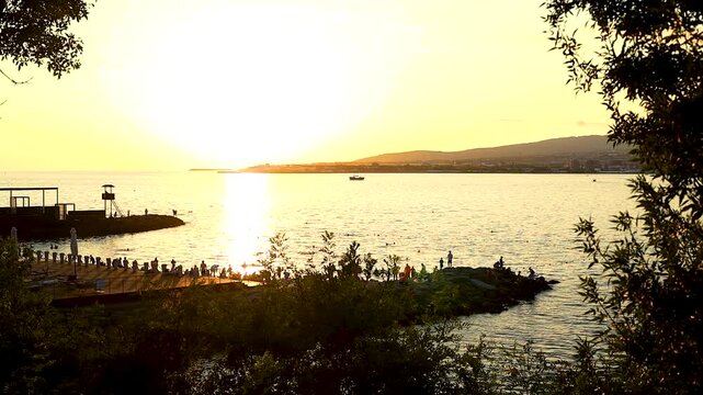 Sunset casts a warm glow over the water, highlighting silhouettes of people along the shore and boats in the distance, framed by greenery