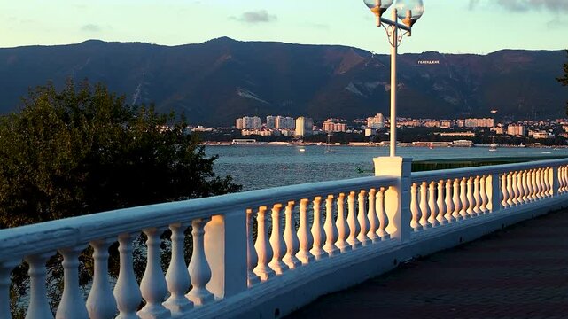 Scenic coastal promenade with white railing and street lamps overlooking the bay, mountains in the background, and urban skyline at sunset in a tranquil setting
