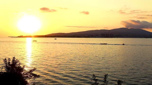 Sunset casts a warm glow over the water, highlighting silhouettes of people along the shore and boats in the distance, framed by greenery
