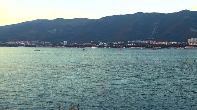 Coastal cityscape features colorful boats on serene water, with mountains rising in the background, capturing the peaceful atmosphere of the shoreline