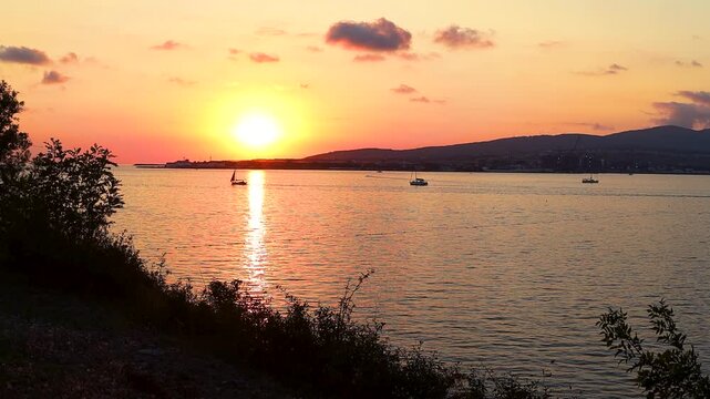 Sunset casts warm hues over water as boats sail peacefully, framed by trees and distant hills, creating a tranquil coastal atmosphere
