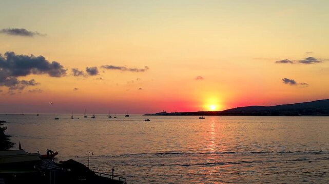 Sunset casts vibrant orange and yellow colors over the ocean, with boats gently bobbing on the water's surface, creating a serene evening atmosphere