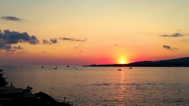 Sunset casts vibrant orange and yellow colors over the ocean, with boats gently bobbing on the water's surface, creating a serene evening atmosphere