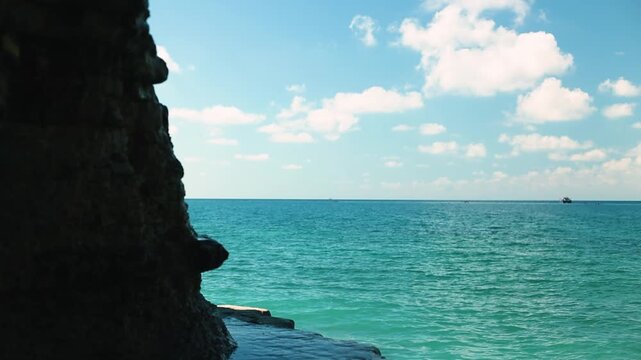 Coastal scene featuring turquoise ocean waves meeting rocky shore under a clear blue sky with fluffy clouds and a distant boat on the horizon