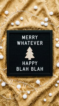 Black letter board with merry whatever text and a white christmas tree icon on a golden fabric background with small white decorative balls for a funny holiday greeting card