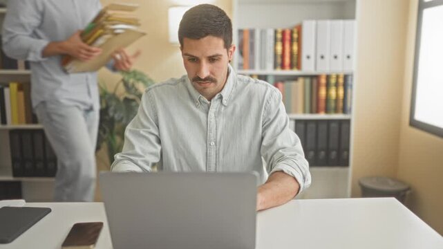 Man at laptop looks up while partner places a stack of folders on the desk in an office; workload concern collaboration.