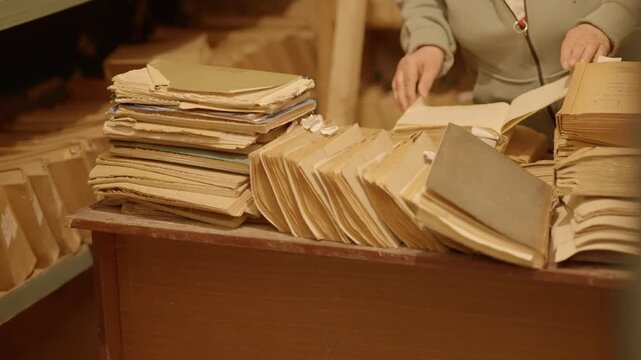 Archivist Reviewing Stacks Of Old Documents And Folders On Wooden Desk In Storage Room