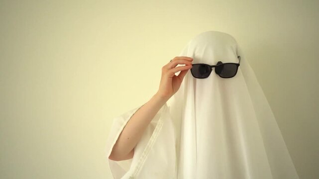 White bedsheet ghost adjusting black sunglasses on the head. Halloween costume concept, white sheet, spooky character, playful fashion and mystery, 4k.
