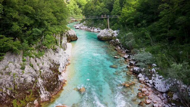 Aerial drone view of the turquoise river running between rocky banks with a small suspension bridge crossing it downstream, Soca Valley, Slovenia