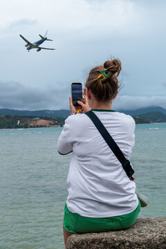 Tourist photographing an airplane flying over a tropical coastal destination, representing travel anticipation, tourism, vacation, air travel and the excitement of a journey.