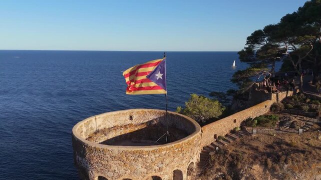 The flag of Catalonia flies on the tower of a fortress on the seashore at sunset on a sunny day in the tourist town of Tossa De Mar, Catalonia, Spain