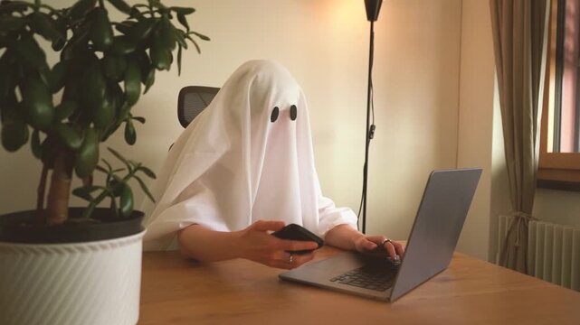 Funny ghost working from home, checking the phone beside an open laptop at a worm autumn office on Halloween