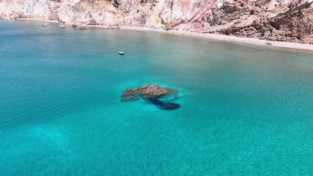 Firiplaka Beach curves beneath layered red, white and ochre volcanic cliffs, with a small boat over vivid turquoise water along the southern coast of Milos, Cyclades, Greece, drone forward flyover
