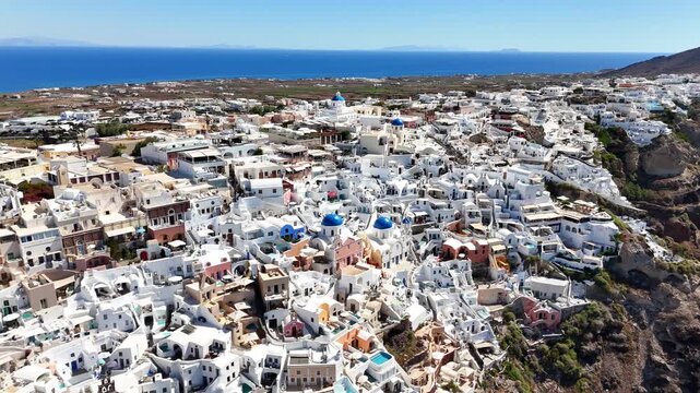 Oia village spreads across the caldera rim with the blue domes of Agios Spyridon and Anastasi among dense whitewashed buildings, Santorini, Greece, static drone orbit shot