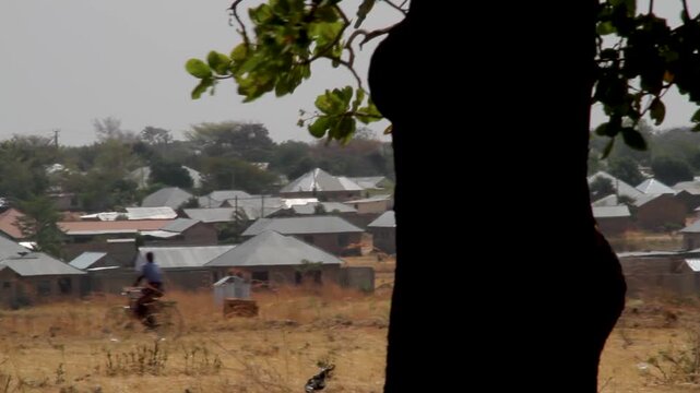 A scenic view of a small village in a rural area, featuring simple houses with metal roofs set against a backdrop of trees and dry, golden-brown grass.
