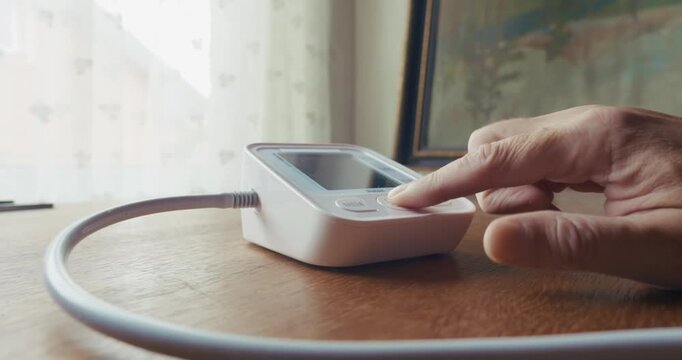 Home patient picks up a blood pressure armband from a table and activates the monitor for self-testing. Illustrates daily hypertension management, cardiovascular health, and home medical routines.