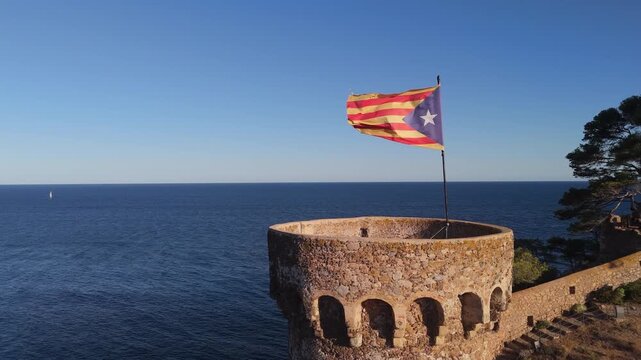 The flag of Catalonia flies on the tower of a fortress on the seashore at sunset on a sunny day in the tourist town of Tossa De Mar, Catalonia, Spain