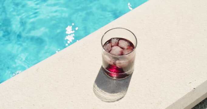 Refreshing cold red drink with ice placed beside a swimming pool on a sunny summer day, representing relaxation, vacation and poolside refreshment.