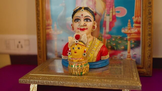 Colorful South Indian wedding doll idol sits before a framed Hindu deity poster on a decorated platform.