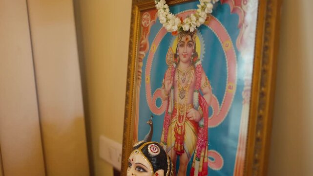 Colorful South Indian Gauri doll sits before a framed Hindu deity portrait decorated with a flower garland at a wedding.